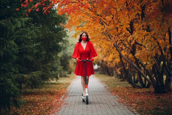 Молода жінка з електричним скутером у червоній сукні в осінньому міському парку — стокове фото