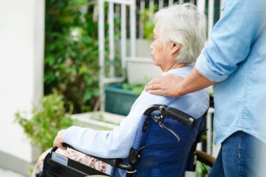 Bakıcı hastanede tekerlekli sandalyede oturan Asyalı yaşlı kadın maluliyet hastasına yardım ediyor..