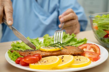 Hastanede sağlıklı yiyecekler için somon kazığı ve sebze salatası yiyen Asyalı yaşlı kadın hasta..