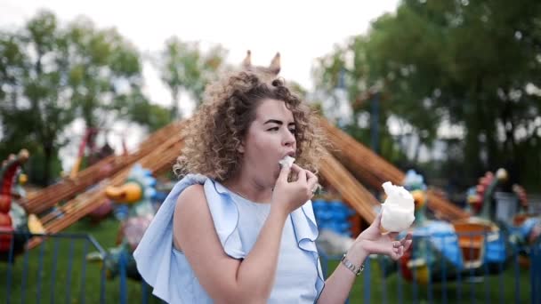 Vídeo retrato de mulher bonita com cabelo encaracolado come algodão doce e sorri no parque de diversões no verão, carrosséis e atrações em lunapark, entretenimento e descanso nos fins de semana de verão — Vídeo de Stock