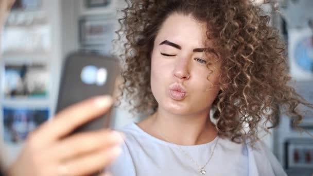 Видео портрет, как кудрявая красивая молодая женщина улыбается в камеру и делает селфи с ее телефоном в различных позах, извращенная девушка делает фотографии в социальных сетях, фотографии после того, как делает прическу — стоковое видео