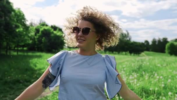 Kinky γυναίκα τινάζει τις τρίχες με τον άνεμο και χαμόγελα στο καταπράσινο κήπο, στην ηλιόλουστη καλοκαιρινή ημέρα, ελκυστικό κορίτσι με τα γυαλιά ηλίου, καλοκαιρινή βόλτα στο πάρκο — Αρχείο Βίντεο