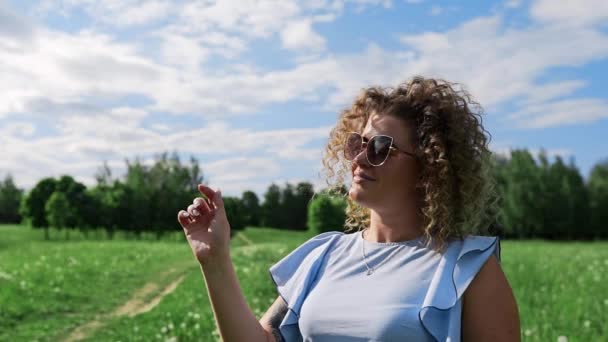 Kinky γυναίκα τινάζει τις τρίχες με τον άνεμο και χαμόγελα στο καταπράσινο κήπο, στην ηλιόλουστη καλοκαιρινή ημέρα, ελκυστικό κορίτσι με τα γυαλιά ηλίου, καλοκαιρινή βόλτα στο πάρκο — Αρχείο Βίντεο