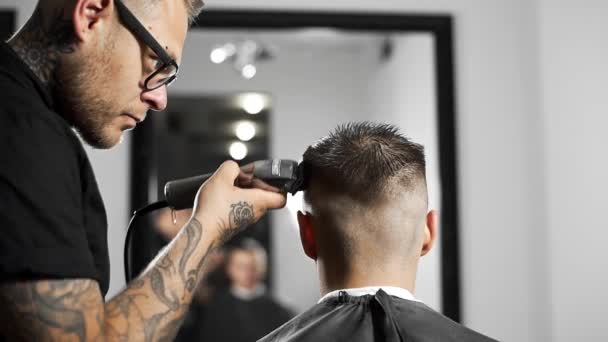 Tattoed barber makes haircut for customer at the barber shop by using hairclipper, mans haircut and shaving at the hairdresser, barber shop and shaving salon — Stock Video