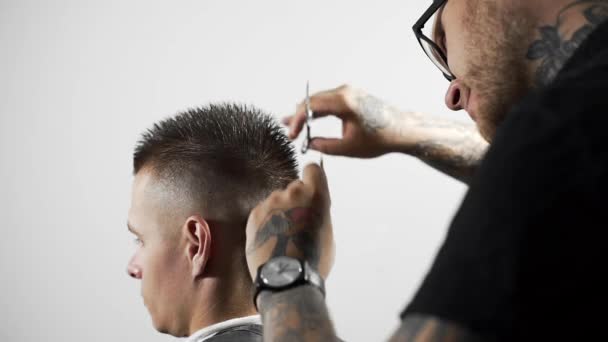 Barbeiro tatuado faz corte de cabelo para o cliente na barbearia usando tesoura e pente, corte de cabelo homem e barbear no cabeleireiro, barbearia e salão de barbear — Vídeo de Stock