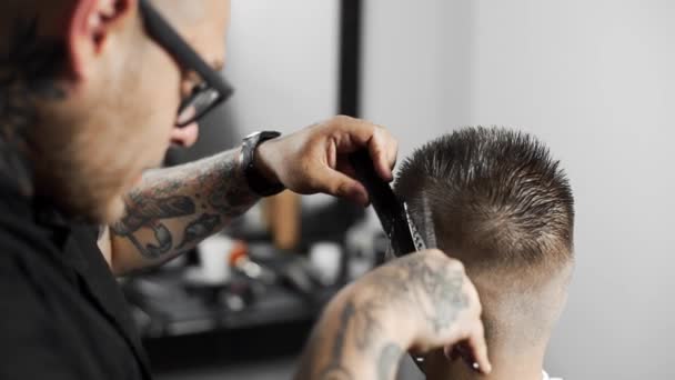 Barbeiro tatuado faz corte de cabelo para o cliente na barbearia usando tesoura e pente, corte de cabelo homem e barbear no cabeleireiro, barbearia e salão de barbear — Vídeo de Stock