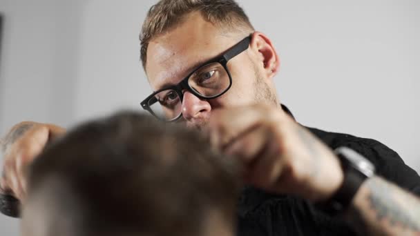 Tattoed barber makes haircut for customer at the barber shop by using scissors and comb , mans haircut and shaving at the hairdresser, barber shop and shaving salon — Stock Video