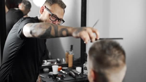 Barbeiro tatuado faz corte de cabelo para o cliente na barbearia, homem corte de cabelo e barbear no cabeleireiro, barbeiro termina o corte de cabelo para o cliente usando pente — Vídeo de Stock