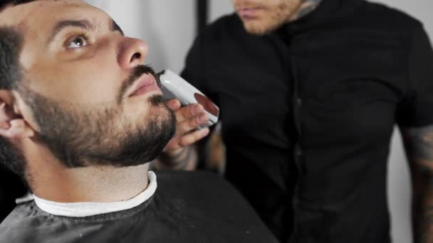 Tattoed barbiere cesoia i clienti barba utilizzando trimmer presso il negozio di barbiere, taglio di capelli mans e rasatura presso il parrucchiere, barbiere e salone da barba — Video Stock