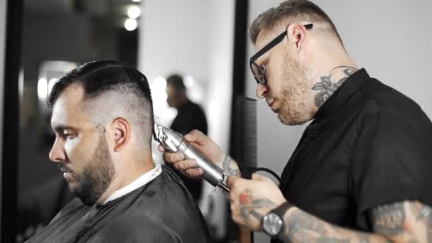 Le coiffeur tatoué fait une coupe de cheveux pour le client au salon de coiffure en utilisant une tondeuse, une coupe de cheveux et un rasage au salon de coiffure, au salon de coiffure et au salon de rasage. — Video
