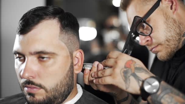 Barbeiro tatuado faz corte de cabelo para o cliente na barbearia usando hairclipper, corte de cabelo homem e barbear no cabeleireiro, barbearia e salão de barbear — Vídeo de Stock