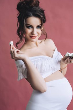 Seksi hamile anne, şirin göbek, gelecekteki parrents portresi beyaz elbise gülümsüyor ve yiyecek Marshmallow, uzun kahverengi saçlı güzel hamile model