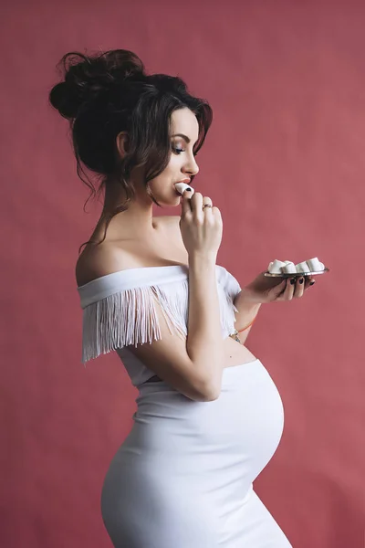 美丽的怀孕模型与长棕色头发在白色礼服微笑和吃棉花糖, 性感怀孕母亲的画像, 可爱的腹部, 未来 parrents 下班 — 图库照片