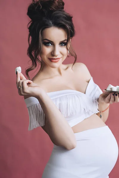Mooie zwangere model met lang bruin haar in de witte jurk glimlacht en eet marshmallows, portret van sexy zwangere moeder, schattige buik, toekomstige parrents — Stockfoto