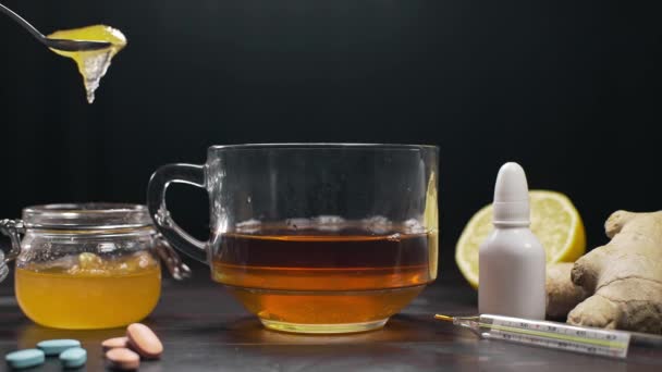 La miel se añade a la copa de vidrio con té negro, té de limón y la miel está sobre la mesa con pastillas y termómetro, tratamiento de vitaminas naturales contra la enfermedad y la enfermedad, coger un resfriado, inmunidad — Vídeos de Stock