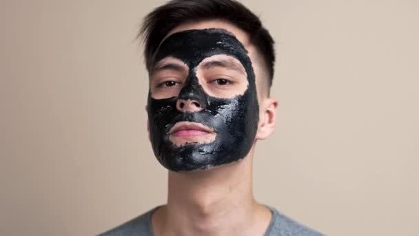 Красивый молодой парень в черной косметической маске смотрит в камеру в замедленной съемке, видео портрет, уход за лицом, селфи видео, красотка блоггер, косметика для лица — стоковое видео