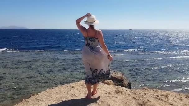 Mujer de pie en la colina en el lado del mar en un día soleado y ventoso con la celebración de su sombrero contra el viento, vacaciones en el mar, recuerdos de verano, relajarse en la playa, el mar y la recreación, arena y agua azul — Vídeos de Stock
