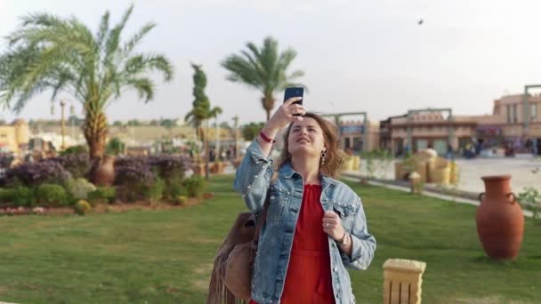 Mujer de camisa roja y chaqueta jeans hace fotos móviles de los lugares de interés, turismo de vacaciones, fotografía móvil — Vídeos de Stock
