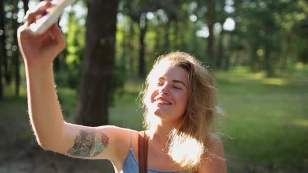 Mujer bastante europea en vestido hace selfie y mensajes de texto con su teléfono móvil en el bosque al atardecer, selfie al aire libre, utilizando el teléfono móvil en el bosque de verano — Vídeos de Stock