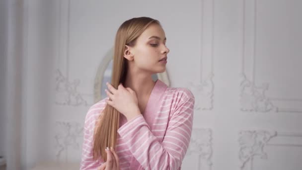 Mujer bonita en albornoz está peinando su cabello largo en el dormitorio de la mañana con luz solar, cuidado del cabello, mañana feliz, comienzo del día, 4k UHD Prores HQ 422 — Vídeos de Stock