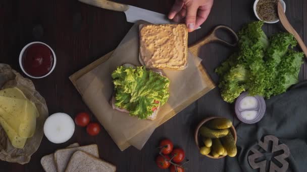 Letak roti lapis yang datar dengan ham dan salad di papan kayu dalam berkas cahaya, koki tangan menyelesaikan roti lapis, membuat makanan cepat saji di dapur, Full HD Prores HQ 422 — Stok Video