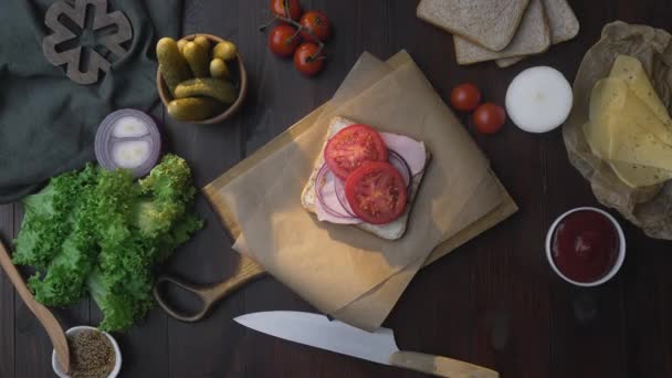 La disposición plana del sándwich con jamón en rodajas y verduras en el tablero de madera en el haz de luz, la mano de los chefs agrega encurtidos deslizados al sándwich, haciendo de la comida rápida en la cocina, Full HD Prores — Vídeos de Stock