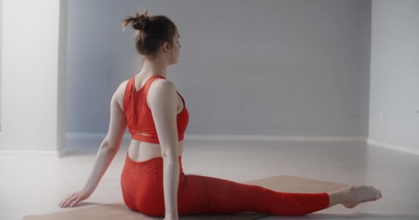 Jeune jolie femme en survêtement de sport rouge s'assoit sur la ficelle et démontre des exercices d'étirement et de yoga au ralenti, la personne fait de l'entraînement physique, de l'étirement et de la gymnastique, 4k 120p Prores HQ — Video
