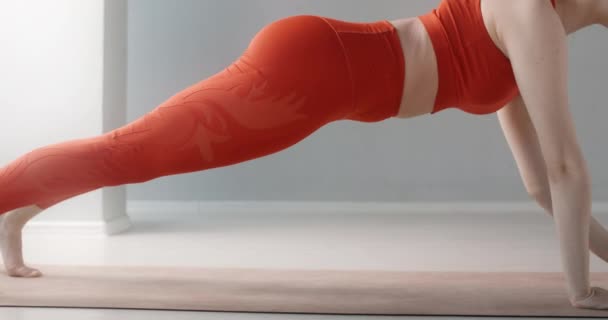 Joven bonita mujer en chándal rojo hace ejercicio de "tablón" en el suelo en el estudio de yoga, persona hace entrenamiento físico, conceptos básicos de yoga, estiramiento y gimnasia, 4k 120p Prores HQ 10 bit — Vídeos de Stock