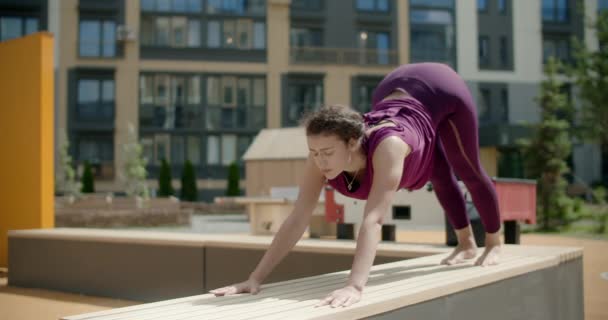 Жінка розтягує і робить вправи з йоги на лавці на подвір'ї житлового будинку, фітнес-класу в міському середовищі, класу йоги на відкритому повітрі, фізичного навантаження в місті, 4k 120p Prores HQ — стокове відео