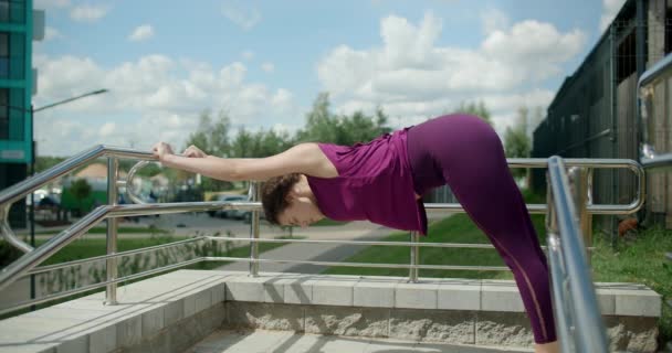 Femme athlétique fait des exercices d'étirement penché sur les mains courantes dans la cour d'un complexe résidentiel appartement, remise en forme dans l'environnement urbain, cours de yoga en plein air, entraînement physique dans la ville, 4k — Video