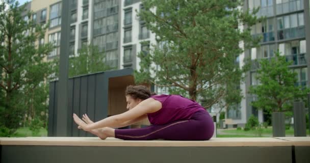 Kadın bir apartmanın bahçesindeki bankta ayaklarını öne eğiyor, şehir ortamında spor yapıyor, yoga sınıfında açık havada, şehirde fiziksel egzersiz yapıyor, 4k 120p Prores karargahında. — Stok video