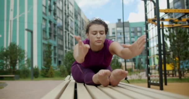 Mujer dobla hacia adelante a sus pies en el banco en el patio de un edificio de apartamentos, fitness en el entorno urbano, clases de yoga al aire libre, entrenamiento físico en la ciudad, 4k 120p HQ de Prores — Vídeo de stock