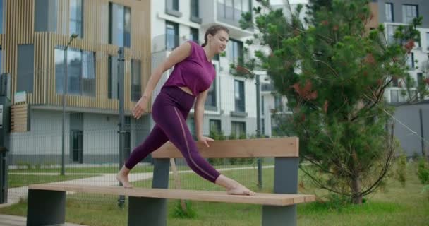 Atletik kadın, bir apartmanın bahçesindeki bankta sicim üzerinde oturuyor, şehir ortamında fitness, yoga sınıfı açık havada, şehirde fiziksel egzersiz, 4k 120p Prores HQ — Stok video