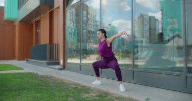 Atletik kadın, cam bina cephesi yakınındaki çimlerde spor yapıyor şehir ortamında spor yapıyor, yoga sınıfında açık havada, şehirde fiziksel antrenman yapıyor, 4k 120p Prores.