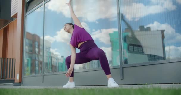 Atletische vrouw doet fitnessoefeningen op het gras in de buurt van glazen gebouw gevel van appartementencomplex, fitness in de stedelijke omgeving, yoga klasse buiten, fysieke training in de stad, 4k 120p Prores — Stockvideo