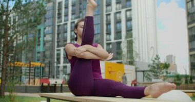 Kadın apartmanın bahçesindeki bankta bacaklar için esneme ve yoga egzersizleri yapıyor. Şehir ortamında fitness, yoga sınıfı açık havada, şehirde fiziksel egzersiz, 4k 120p Prores.