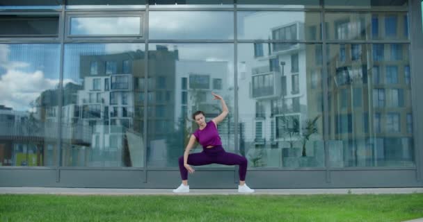 Mujer atlética hace ejercicios de fitness en la hierba cerca de la fachada del edificio de vidrio del complejo de apartamentos, fitness en el entorno urbano, clases de yoga al aire libre, entrenamiento físico en la ciudad, 4k 120p Prores — Vídeos de Stock