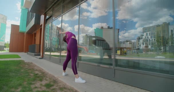 Mujer atlética hace ejercicios de fitness frente a la fachada del edificio de vidrio del complejo de apartamentos, fitness en el entorno urbano, clases de yoga al aire libre, entrenamiento físico en la ciudad, 4k 120p HQ de Prores — Vídeo de stock