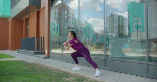 Mujer atlética hace ejercicios de fitness en la hierba cerca de la fachada del edificio de vidrio del complejo de apartamentos, fitness en el entorno urbano, clases de yoga al aire libre, entrenamiento físico en la ciudad, 4k 120p Prores — Vídeos de Stock