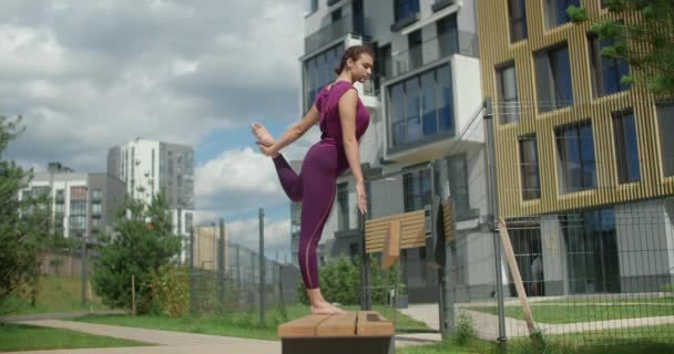 Спортивная женщина делает вертикальную бечевку стоя на скамейке, упражнения на растяжку в городской среде, йога класс на открытом воздухе, физические тренировки и фитнес в городе, 4k 120p Прорес HQ — стоковое видео