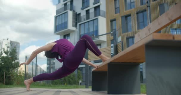 Atletische vrouw doet twine op de bank in een openbare tuin, stretching oefeningen in de stedelijke omgeving, yoga klasse buiten in slow motion, fysieke training en fitness in de stad, 4k 120p Prores HQ — Stockvideo