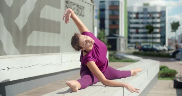 Atletische vrouw zit op een touw op de bank en doet dends aan de zijkanten, stretching in de tuin van een appartementencomplex, fitness in de stedelijke omgeving, yoga buiten, fysieke training in de stad — Stockvideo