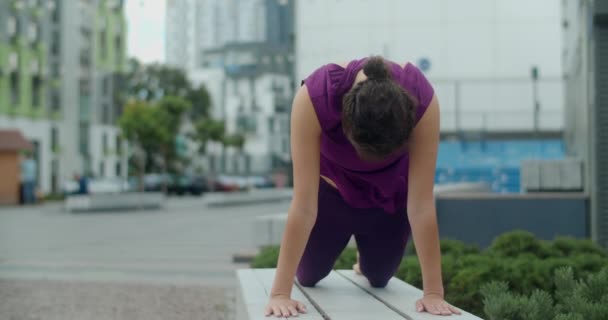 Vrouw doet stretching en yoga oefeningen op de bank in de tuin van een appartementencomplex, fitness in de stedelijke omgeving, yoga klasse buiten, fysieke training in de stad, 4k 60p Prores HQ — Stockvideo