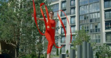 Kırmızılı genç kadın jimnastikçi apartman bahçesinde kurdele ile spor yapıyor, jimnastikçi rezidans alanında akrobatik egzersizler yapıyor, şehir ortamında fitness, 4k 120p Prores karargahı