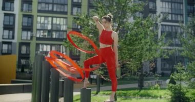 Kırmızılı genç kadın jimnastikçi apartman bahçesinde kurdele ile spor yapıyor, jimnastikçi rezidans alanında akrobatik egzersizler yapıyor, şehir ortamında fitness, 4k 120p Prores karargahı
