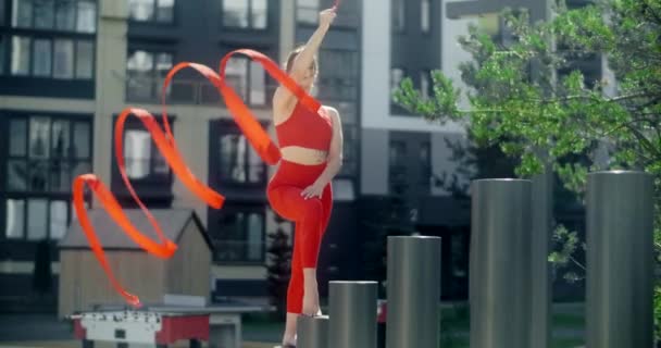 Jonge vrouw in het rood voert callisthenics met lint in de libing appartement werf, gymnast doet acrobatische oefeningen in een residentiële ruimte, fitness in de stedelijke omgeving, 4k 120p Prores HQ — Stockvideo