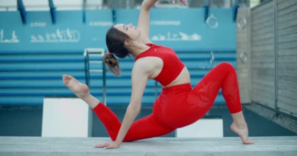 Kobieta w czerwonym stroju sportowym wykonuje ćwiczenia gimnastyczne na ławce na placu zabaw na świeżym powietrzu, fitness w środowisku miejskim, zajęcia jogi, ćwiczenia fizyczne w mieście, 4k 120p Prores HQ — Wideo stockowe