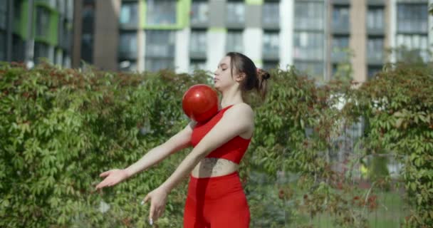 Žena v červeném sportovním kostýmu provádí kallisthenická cvičení s gymnastickým míčkem venku, sport v městském prostředí, gymnastika ve městě, 4k 120p Prores HQ — Stock video