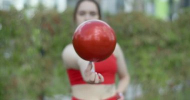 Kırmızı spor kostümlü bir kadın parmağında jimnastik topu, açık şehir ortamında spor, şehirde jimnastik, 4K 120p Prores HQ