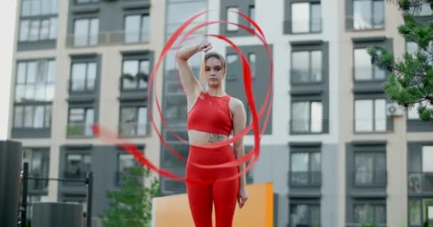 Jonge vrouw in het rood voert callisthenics met lint in de libing appartement werf, gymnast doet acrobatische oefeningen in een residentiële ruimte, fitness in de stedelijke omgeving, 4k 120p Prores HQ — Stockvideo